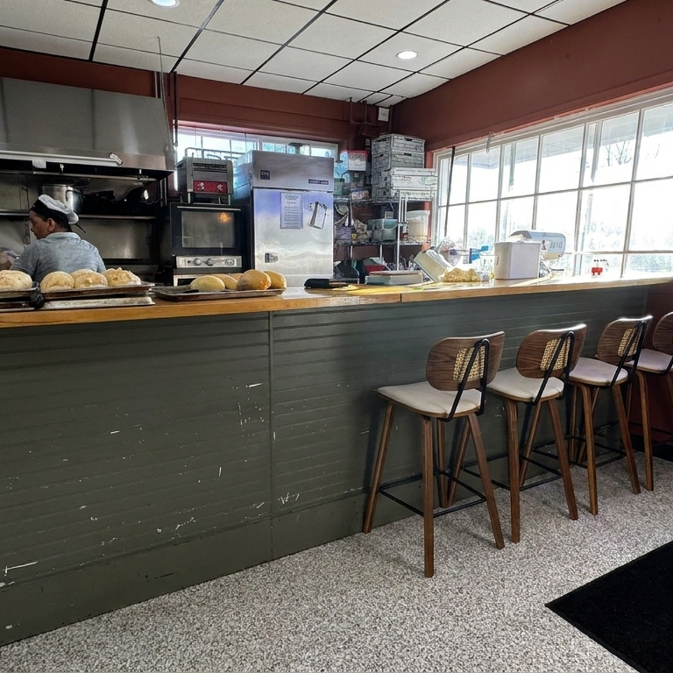 Bakery counter with fresh loaves, commercial kitchen equipment, and four bar stools beside bright front windows.