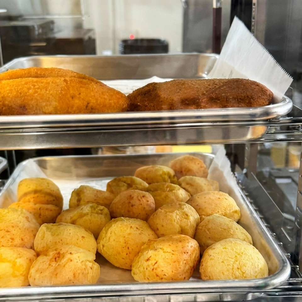 Trays of golden baked pastries and round cheese breads displayed on warming racks behind a glass counter.